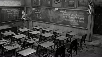 Movie still from “Blackboard Jungle” (1955), directed by Richard Brooks – A man standing in front of a chalkboard with a bunch of desks; Wide shot, High angle