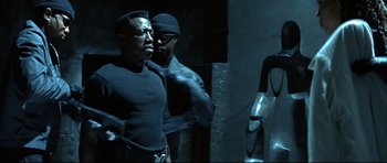 Movie still from “Blade” (1998), directed by Stephen Norrington – A man in a black shirt is standing next to another man in a black hat; Medium shot, Over the shoulder angle