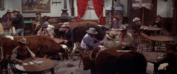 Movie still from “Blazing Saddles” (1974), directed by Mel Brooks – A group of men sitting around a cow in a room; Wide shot, High angle