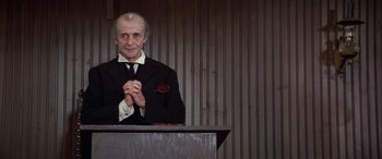 Movie still from “Blazing Saddles” (1974), directed by Mel Brooks – An older man in a black suit clasps his hands while standing at a podium; Medium shot, Low angle