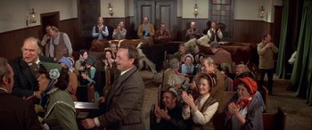 Movie still from “Blazing Saddles” (1974), directed by Mel Brooks – A group of people sitting in a room with cows in the background; Wide shot, High angle