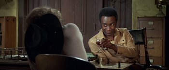 Movie still from “Blazing Saddles” (1974), directed by Mel Brooks – A man sitting at a chess table holding a chess piece; Close Up shot, Over the shoulder angle