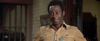 Movie still from “Blazing Saddles” (1974), directed by Mel Brooks – A man in a yellow and brown shirt is smiling for the camera; Close Up shot, Over the shoulder angle