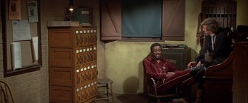 Movie still from “Blazing Saddles” (1974), directed by Mel Brooks – A man sitting in a chair in front of a card catalog; Wide shot, High angle