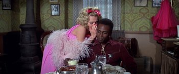 Movie still from “Blazing Saddles” (1974), directed by Mel Brooks – A woman in a pink dress whispering to a man in a maroon shirt; Medium shot, Over the shoulder angle