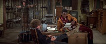 Movie still from “Blazing Saddles” (1974), directed by Mel Brooks – Two men sitting at a table in front of an open fireplace; Medium shot, High angle