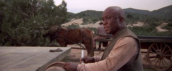 Movie still from “Blazing Saddles” (1974), directed by Mel Brooks – A man sitting next to a horse on a dirt field; Medium shot, Low angle