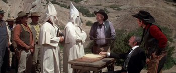 Movie still from “Blazing Saddles” (1974), directed by Mel Brooks – A group of men in white clothes and black hats standing next to a table; Medium shot, High angle