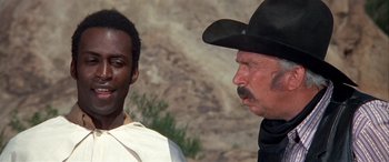 Movie still from “Blazing Saddles” (1974), directed by Mel Brooks – A man and a man in a cowboy hat talking to each other; Close Up shot, Over the shoulder angle