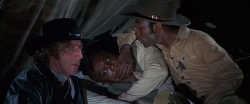 Movie still from “Blazing Saddles” (1974), directed by Mel Brooks – Two men in hats and one is laying on the ground and the other is leaning on another man; Close Up shot, Over the shoulder angle