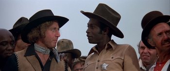 Movie still from “Blazing Saddles” (1974), directed by Mel Brooks – Two men in cowboy hats and brown jackets talking to each other; Close Up shot, Low angle