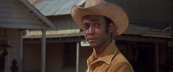 Movie still from “Blazing Saddles” (1974), directed by Mel Brooks – A man wearing a cowboy hat in front of a building; Close Up shot, Over the shoulder angle