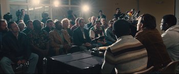 Movie still from “Bleed for This” (2016), directed by Ben Younger – A group of people sitting in front of a microphone in a room; Medium shot, Over the shoulder angle