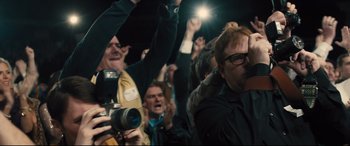Movie still from “Bleed for This” (2016), directed by Ben Younger – A group of people are gathered in a crowd and one person is holding a camera; Medium shot, Over the shoulder angle