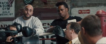 Movie still from “Bleed for This” (2016), directed by Ben Younger – Three men in a boxing ring with one wearing a black shirt and one wearing a white shirt; Medium shot, Over the shoulder angle