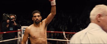 Movie still from “Bleed for This” (2016), directed by Ben Younger – A man in a boxing ring raises his arm in the air; Medium shot, Over the shoulder angle