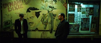 Movie still from “Bleeder” (1999), directed by Nicolas Winding Refn – A man standing in front of a painting of a motorcycle; Medium shot, Over the shoulder angle