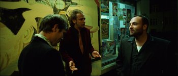 Movie still from “Bleeder” (1999), directed by Nicolas Winding Refn – A man in a red jacket and a tie standing in front of two other men; Medium shot, Over the shoulder angle