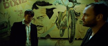Movie still from “Bleeder” (1999), directed by Nicolas Winding Refn – A man standing in front of a painting of a man on a motorcycle; Medium shot, Low angle