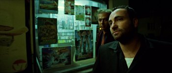 Movie still from “Bleeder” (1999), directed by Nicolas Winding Refn – Two men standing next to each other in front of movie posters; Medium shot, Low angle