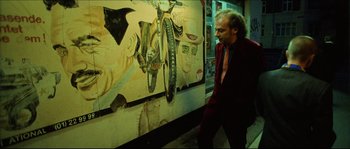 Movie still from “Bleeder” (1999), directed by Nicolas Winding Refn – A man in a red suit walking past a painting of a bicycle; Medium shot, Low angle