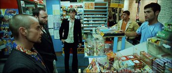 Movie still from “Bleeder” (1999), directed by Nicolas Winding Refn – A man standing in front of a store filled with lots of items; Medium shot, Over the shoulder angle