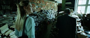 Movie still from “Bleeder” (1999), directed by Nicolas Winding Refn – A woman standing in front of stacks of books in a room; Medium shot, Low angle