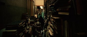 Movie still from “Bleeder” (1999), directed by Nicolas Winding Refn – A woman standing in front of stacks of books in a room; Wide shot, High angle