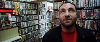 Movie still from “Bleeder” (1999), directed by Nicolas Winding Refn – A man standing in front of a room filled with lots of movies; Close Up shot, Low angle