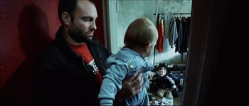 Movie still from “Bleeder” (1999), directed by Nicolas Winding Refn – A man holding a child in his arms in a room; Medium shot, Over the shoulder angle