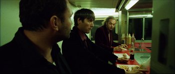 Movie still from “Bleeder” (1999), directed by Nicolas Winding Refn – A group of men sitting at a table eating pizza; Medium shot, Over the shoulder angle