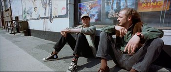 Movie still from “Bleeder” (1999), directed by Nicolas Winding Refn – Two young men sitting on the ground in front of a movie poster; Medium shot, Low angle