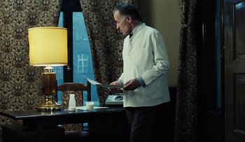 Movie still from “Blind Chance” (1987), directed by Krzysztof Kieslowski – A man standing in front of a table holding papers in his hands; Medium shot, Low angle