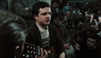 Movie still from “Blind Chance” (1987), directed by Krzysztof Kieslowski – A man playing a guitar in front of a group of people; Close Up shot, High angle