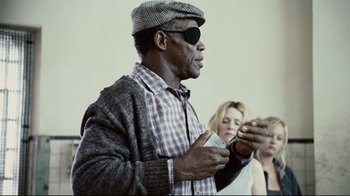 Movie still from “Blindness” (2008), directed by Fernando Meirelles – A man wearing a hat and glasses is holding a cell phone in his hand; Medium shot, Over the shoulder angle