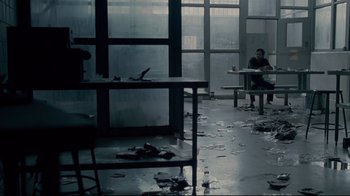 Movie still from “Blindness” (2008), directed by Fernando Meirelles – A person sitting at a table in a room with broken windows; Wide shot, High angle