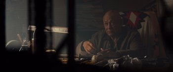 Movie still from “Blood Quantum” (2019), directed by Jeff Barnaby – An older man sitting at a table in a dark room; Medium shot, Over the shoulder angle