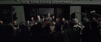 Movie still from “The Amazing Spider-Man” (2012), directed by Marc Webb – A group of people standing around a building with a police officer; Wide shot, Over the shoulder angle