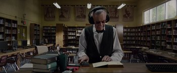 Movie still from “The Amazing Spider-Man” (2012), directed by Marc Webb – An older man wearing headphones while reading a book in a library; Medium shot, Low angle