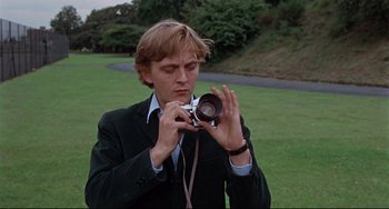 Movie still from “Blow-Up” (1966), directed by Michelangelo Antonioni – A man in a suit taking a picture with a camera; Close Up shot, Low angle