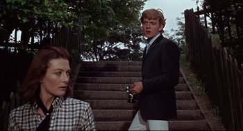 Movie still from “Blow-Up” (1966), directed by Michelangelo Antonioni – A man in a suit and tie standing next to a woman in a checkered shirt; Medium shot, Low angle