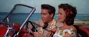 Movie still from “Blue Hawaii” (1961), directed by Norman Taurog – A man and a woman sitting in the drivers seat of a car; Medium shot, Low angle