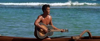 Movie still from “Blue Hawaii” (1961), directed by Norman Taurog – A shirtless man sitting on a dock with a guitar; Medium shot, High angle
