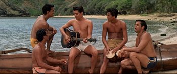 Movie still from “Blue Hawaii” (1961), directed by Norman Taurog – A group of young men sitting next to a body of water; Wide shot, Over the shoulder angle