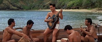Movie still from “Blue Hawaii” (1961), directed by Norman Taurog – A man sitting on a boat playing a guitar; Wide shot, Low angle