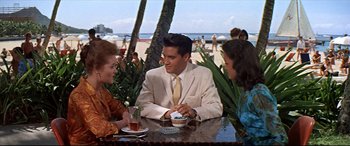 Movie still from “Blue Hawaii” (1961), directed by Norman Taurog – A man sitting at a table in front of two other people; Medium shot, Over the shoulder angle