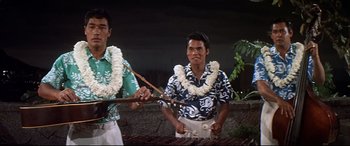 Movie still from “Blue Hawaii” (1961), directed by Norman Taurog – Two men in hawaiian garb are playing a musical instrument; Medium shot, Low angle