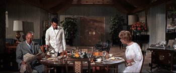 Movie still from “Blue Hawaii” (1961), directed by Norman Taurog – A man standing at a dining room table with plates of food on top of it; Wide shot, High angle