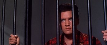 Movie still from “Blue Hawaii” (1961), directed by Norman Taurog – A man in a red shirt behind a metal fence; Close Up shot, Low angle