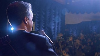 Movie still from “Bob Roberts” (1992), directed by Tim Robbins – A man with a microphone in front of an audience; Medium shot, Low angle